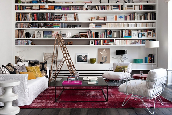Nowoczesna aranżacja salonu z bialymi półkami na ścianie,drabiną,nowoczesnymi metalowymi fotelami w bieli i różowym dywanem vintage