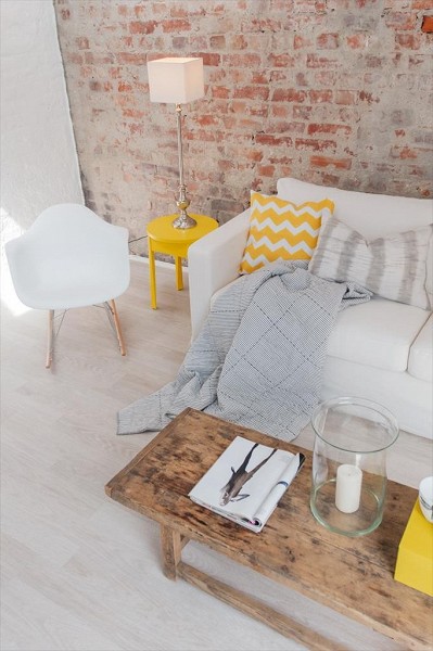 Żółty stolik pomocniczy,poduszki z żółto-białym wzorem szerwonu,drewniany stolik vintage i ściana z cegły