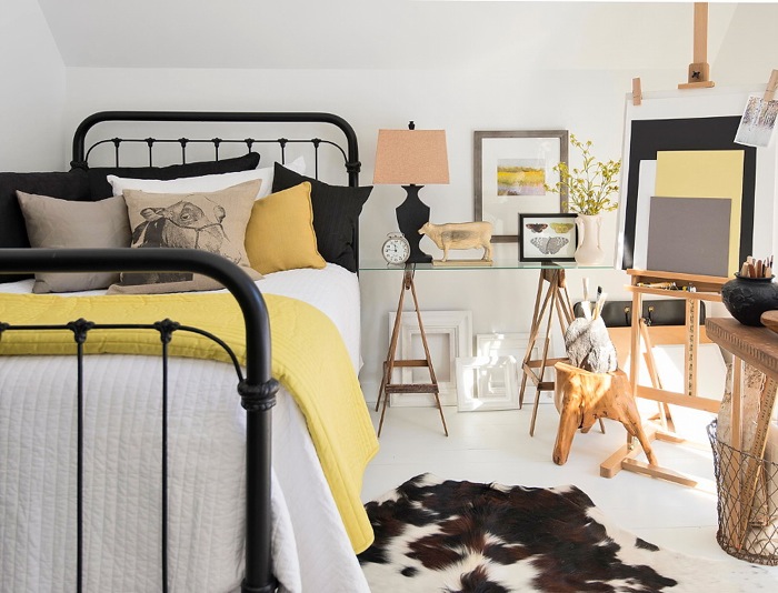 Kute czarne łóżko,stół na drewnianych kozłach ze szklanym blatem,czarno-zółte dekoracje łóżka,czarna lampa z bezowym abażurem i dywan z bydlęcej skóry na białej podłodze