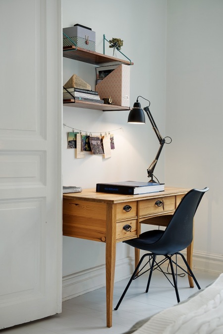 Drewniane miodowe biurko z wiszącymi pólkami na białej scianie w aranżacji mieszkania,czarne krzesło na krzyżakach i czarna lampa biurkowa z przegubami