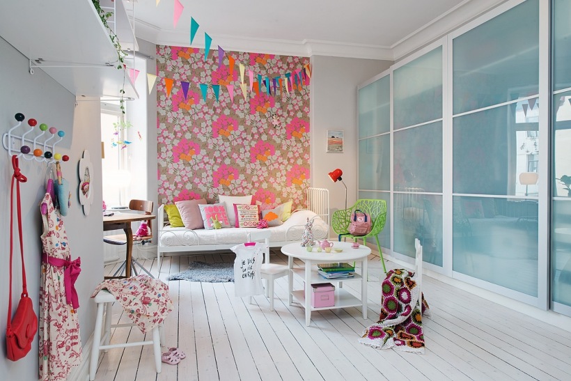 Tapeta ścienna w różowe kwiaty,szafa z przesuwnymi szklanymi i piaskowanymi drzwiami,metalowy wieszak z kolorowymi gałkami i białe mebelki w pokoju dziecięcym