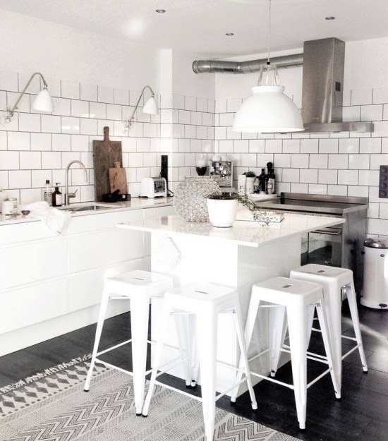 Stalowy okap na białej ścianie z glazurowanych cegiełek w białej kuchni z kuchenną wyspą, białymi stołkami wysokimi i czarną podłogą