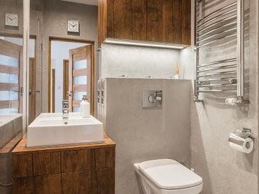 Łazienka w nowoczesnym stylu, w której zestawiono ze sobą szarość oraz ciemne drewno. Wysokie lustro nad umywalką...