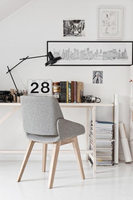 Biały kącik biurowy z nowoczesną czarną lampą, prostym biurkiem,szarym tapicerowanym fotelikiem i biało-czarnymi skandynawskimi grafikami