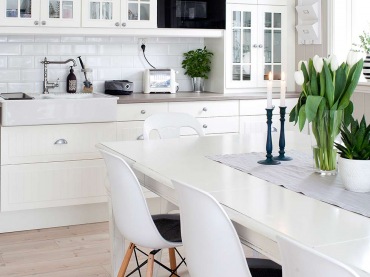 Białe szafki kuchenne zajmują całą ścianę. Tuż przy nich stół i krzesła, również w kolorze białym. To niezwykle jasne i...