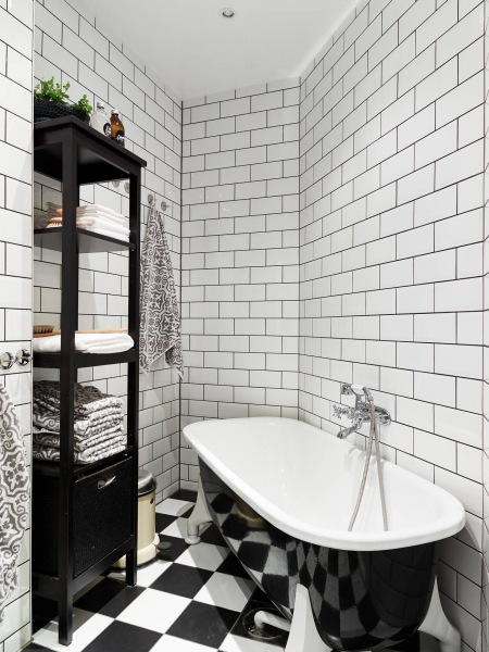 Biała glazura cegiełka na ścianie w łazience, posadzka w szachownicę ,wolnostojaca czarna wanna i czarne etażeki z pólkami