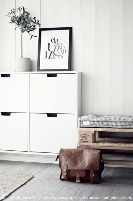Biała wisząca szafka na buty,siedzisko z drewnianej palety,pikowane poduszki w paski,białe panele na ścianie,szara podłoga z desek w przedpokoju w stylu skandynawskim