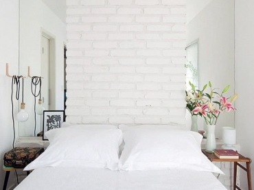 Ściana z białych cegieł za łóżkiem w sypialni nawiązuje do stylu skandynawskiego. Świeże kwiaty w wazonie wprowadzają przyjemny naturalny...