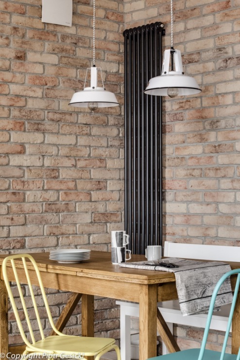 Surowe cegły na ścianie w jadalni wyraźnie eksponują szczególnie kolorowe krzesła przy drewnianym stole. Białe...