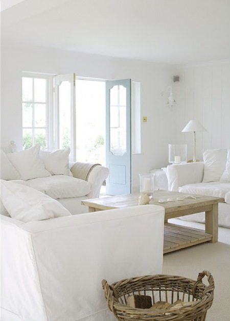 Aranżacja salonu z białymi sofami,drewnianym stolikiem ,wiklinowym koszem i niebieskimi drzwiami