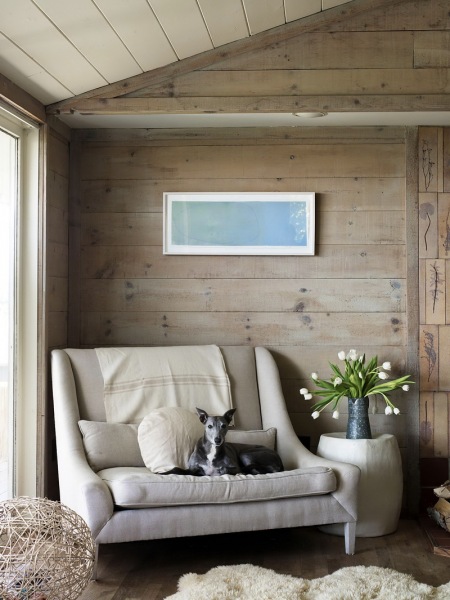 Nowoczesna kanapa w tapicerce złamanej bieli,futrzak i szerokie proste deski na ścianie w aranżacji kącika wypoczynkowego w wakacyjnym domu