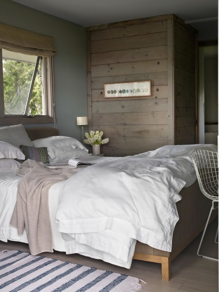Lniana roleta rzymska,drewniane łóżko,szerokie szarawe deski na ścianie i ażurowy biały foteli z metalu