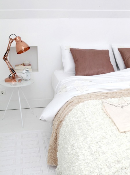 Miedziana lampka i miedziany kolor poduszek w aranżacji białej sypialni