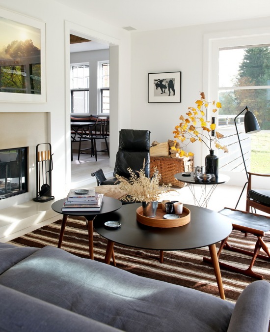 Szara sofa,czarny fotel ze skóry,okrągłe stoliki kawowe w stylu skandynawskim i biało-brązowy dywan w paski w salonie z kominkiem