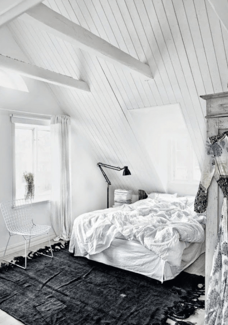 Biała sypialnia ze skośnymi ścianami z białymi belkami i czarnym dywanem z frędzlami na podłodze