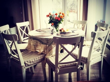 Przyjemna aranżacja jadalni w rustykalnym stylu z okrągłym stołem. Wzorzyste poduszki na krzesłach dekorują przestrzeń,...