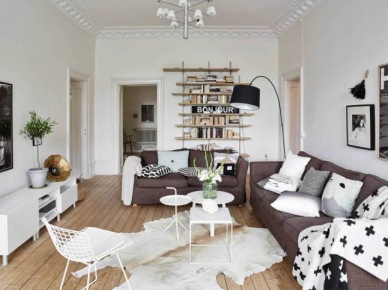 Brązowy narożnik ,druciane białe krzesła,małe biale stoliki okragłe i drewniane pólki na scianie w skandynawskim salonie (25685)