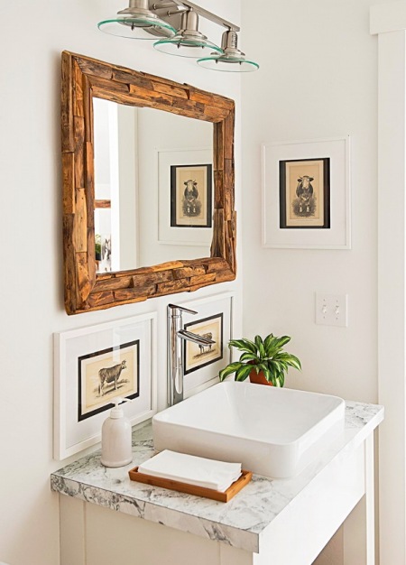 Lustro z drewna,kamienny blat na szafce z umywalką,retro obrazki w białych ramkach na scianie w białej łazience