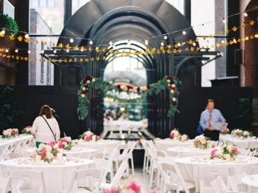 Pomysł na dekoracje sali na wesele. Klasyczne białe obrusy i krzesła, stoły ozdobione bukietami różowych kwiatów, a całość oświetlona girlandami świetlnymi, które znacznie ocieplają surowe...