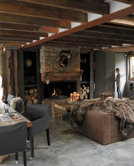 Drewno i naturalny kamień przy kominku w rustykalnym salonie ze skórzaną sofą,futrzakami i betonową podłogą