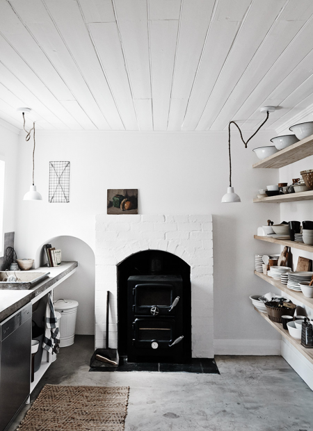 Rustykalny murowany   kominek w białej  kuchni ze ścianą z półkami na kuchenne naczynia i pojemniki