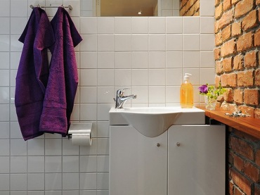 Surowa łazienka, białe kafelki i ciemna podłoga ożywione dzięki ścianie z czerwonej cegły.