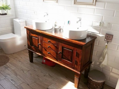 Aranżacja białej łazienki z drewnianą podłogą i rzeźbioną szafką. Dwie umywalki zapewniają wygodny równoległy dostęp dla domowników. Drobne dodatki i białe kafle w bardzo subtelny sposób dekorują łazienkową...