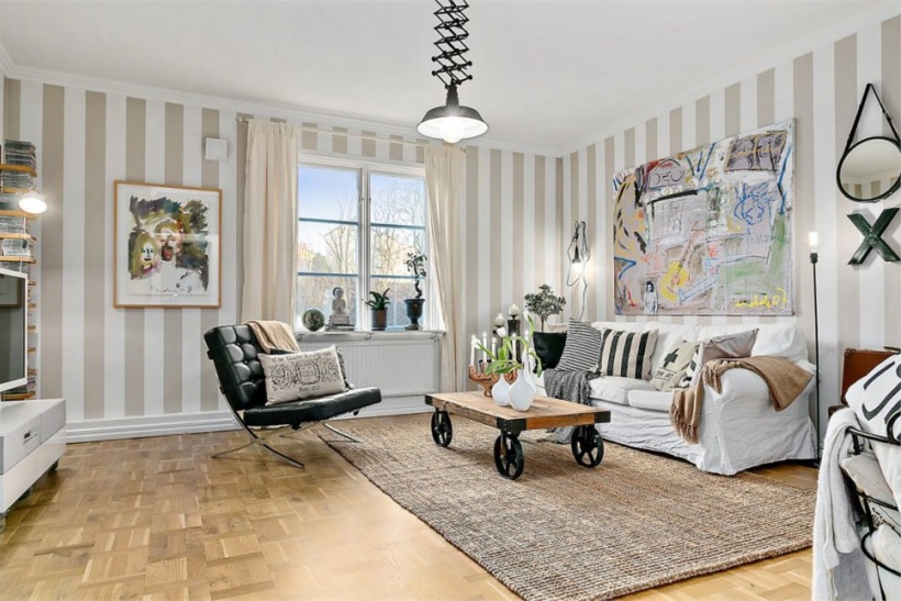 Skandynawski salon z tapetą w beżowe pasy,z biała sofą, jutowym dywanem, czarną lampą i stolikiem na kólkach w stylu industrialnym