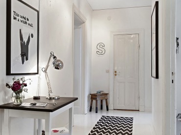 Biały drewniany stół z czarnym blatem,srebrna chromoniklowana lampa biurkowa,czarno-biała grafika,czarno-biały dywan z wzorem szewronu na białej podłodze,drewniany stołek vintage w przedpokoju (25812)