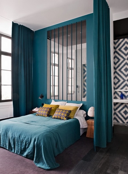 Ciemno-turkusowe zasłony i sciana z lustrzanymi listwami w aranżacji nowoczesnej sypialni z turkusową narzutą,żółtymi poduszkami i grafitową podłogą z desek
