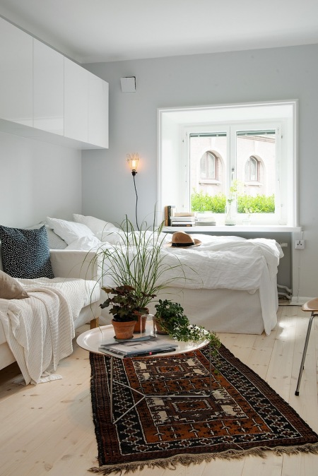 Białe wiszące szafki,łóżko przy oknie i biała sofa, okagłymetalowy stolik z tacą i etniczny brązowy dywan w salonie połączonym z sypialnią