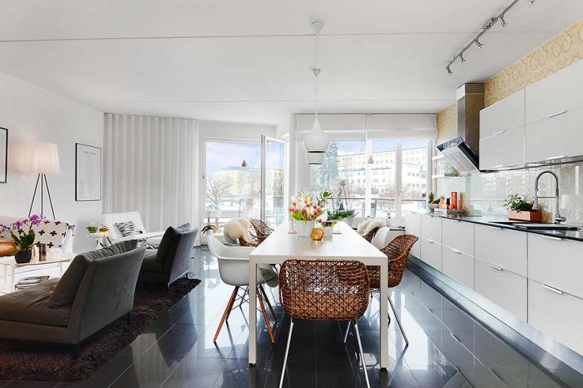 Szare nowoczesne leżanki , biały stół z białymi i miodowymi krzesłami i biała kuchnia w otwartym widoku mieszkania