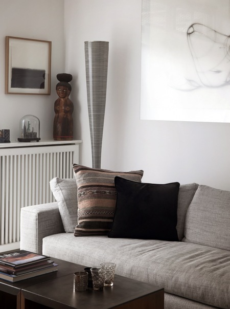 Jasnoszara sofa nowoczesna,brązowo-szara poduszka z etnicznymi wzorami,nowoczesna lampa podłogowa tuba i drewniane posążki