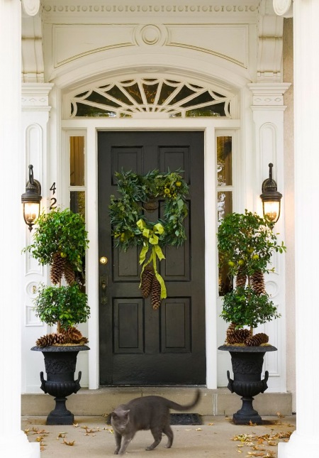 Czarne kinkiety przy czarnych drzwiach z czarnymi gazonami z zielonymi drzewkami i szyszkami i zielony wianek na drzwiach w świątecznej dekoracji wejścia do domu