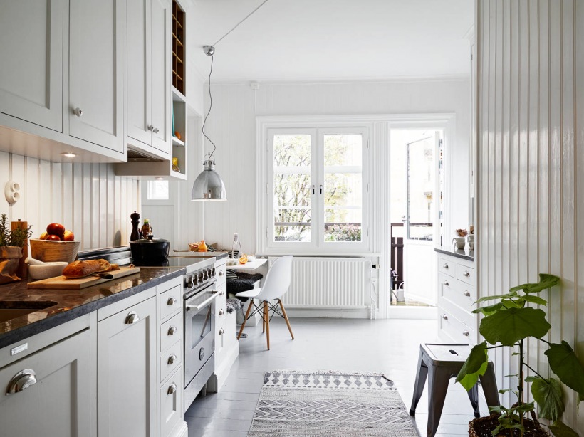 Tradycyjna biała kuchnia skandynawska z zabudowanymi siedziskami, biało-czarnymi poduszkami, srebrną lampą nad stołem i białą boazerią na ścianach