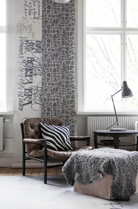 Graficzne wzory tapet w aranzacji skandynawskiego salonu ,czarny drewniany fotel ze skórzana tapicerką,kwadatowy puf z szarym futrzakiem i okragły czarny stolik kawowy