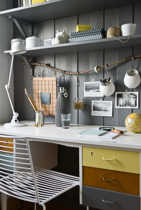 Szare panele na ścianie z półką ,białe biurko z kolorowymi szufladami,białe ażurowe krzesło z metalu i gałąź jako wieszak na biurowe ceramiczne i druciane pojemniki