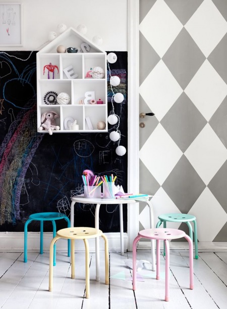 Biało-szara tapeta w romby na drzwiach i czarna ściana z farbą tablicową ,kolorowe metalowe stołki przy okragłym stoliku,półka domek  z bawełnianymi kulami z girlandą