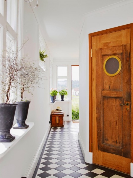 Białe ściany,białoczarna terakota cementowa ułożona w karo,czeresniowe drzwi z okragłym oknem w stylu vintage,drewniana skrzynia,czarne amfory z kamienia