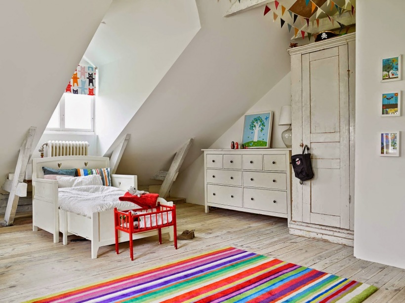 Białe skrzyniowe łóżko dziecięce,biała komoda z szufladami,kolorowa girlanda z proporczykami i kolorowy dywanik w paski w dziecięcym pokoju