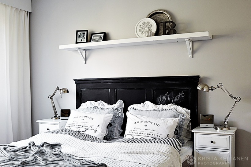 Czarne drewniane wezgłowie łóżka w  białej sypialni,półkanad łóżkiem,metalowe  srebrne lampki nocne,drukowane poduszki na łóżku