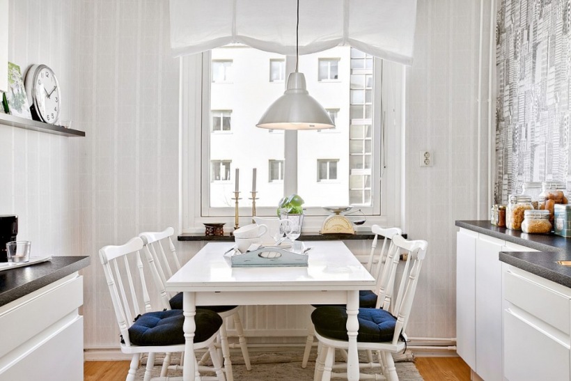 Biała roleta rzymska na oknie w kuchni,białe krzesła z drewena z siedziskami i biały prostokatny stół w stylu skandynawskim