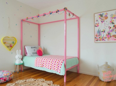 Sypialnia idealna dla dziewczynki: różowe łóżko z baldachimem i dekoracjami w postaci pastelowego nakrycia i poduszek. Na ścianie obraz przedstawiający kwiaty i żółta półeczka w kształcie serca. Przed łóżkiem małej księżniczki pleciony dywanik przypominający kształtem szydełkową...