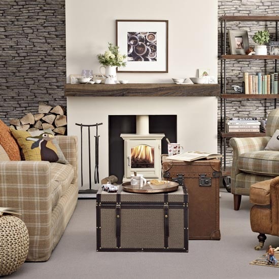 Kamień łupkowy na ścianie w salonie z białym kominkiem z drewnianą półką,kufry skrzynie w roli stolków i kraciaste sofy i fotele