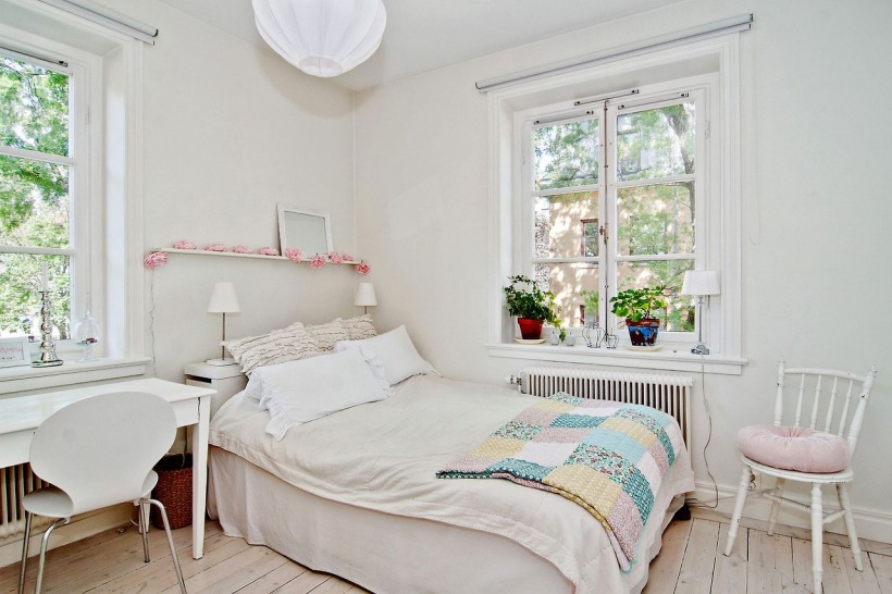 Biała sypialnia z patchworkowa narzuta w pastelach i różową girlandą na małej półce nad łóżkiem