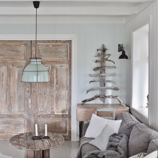 Bielone drzwi z drewna,stół szpula z recyklingu,turkusowa metalowa lampa,szara sofa i świąteczne drzewko z korzeni drzew
