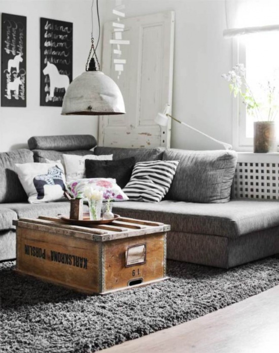 Drewniana skrzynia w roli stolika w aranżacji skandynawskiego salonu z szarą sofą, białą lampa industrialną i biało-czarnymi dekoracjami