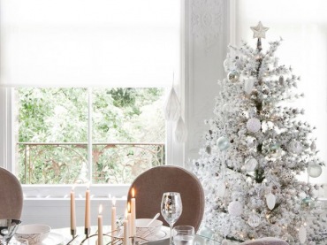 Biała i oszroniona dekoracja zielonej choinki przy okrągłym szklanym stole i tapicerowanymi krzesłami w jadalni z białymi roletami na oknach (27423)
