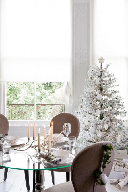 Biała i oszroniona dekoracja zielonej choinki przy okrągłym szklanym stole i tapicerowanymi krzesłami w jadalni z białymi roletami na oknach