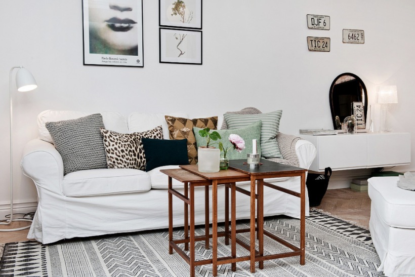 Skandynawskie grafiki na scianie,biała sofa,drewniane stoliki kawowe i biało-szary dywan z etnicznymi wzorami